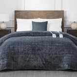 A bed features a herringbone-patterned dark gray heated blanket with dual controllers, white pillows, and a dark gray accent pillow. The bed has a wooden headboard and is flanked by two bedside tables with lamps.