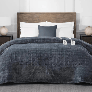 A bed features a herringbone-patterned dark gray heated blanket with dual controllers, white pillows, and a dark gray accent pillow. The bed has a wooden headboard and is flanked by two bedside tables with lamps.