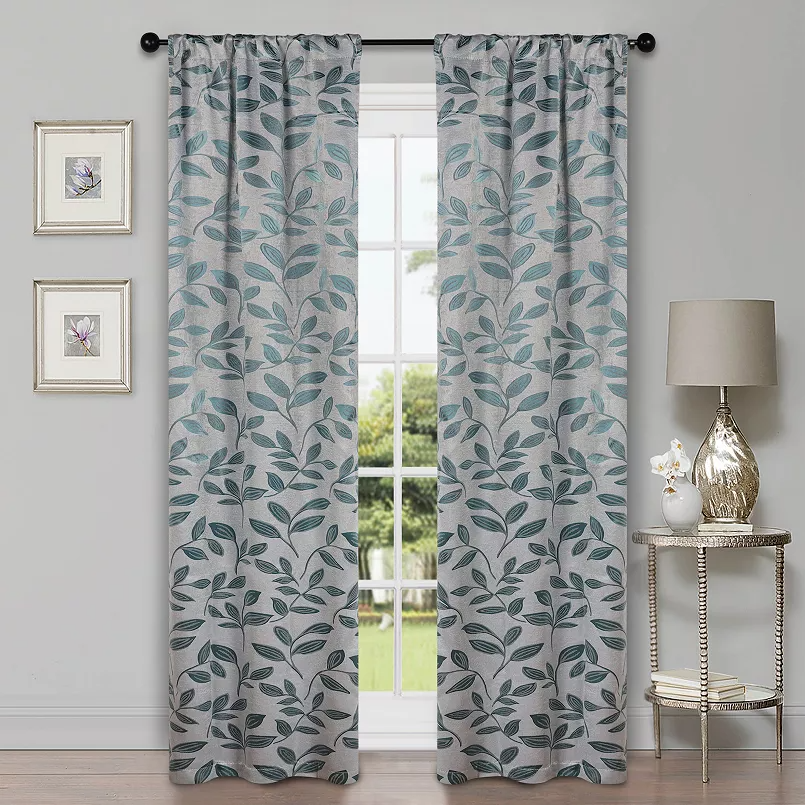 Two light gray curtains with a leaf pattern hang in front of a window. They are accompanied by framed floral artwork on the wall and a lamp on a small decorative table.