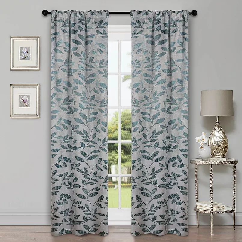 Two light gray curtains with a leaf pattern hang in front of a window. They are accompanied by framed floral artwork on the wall and a lamp on a small decorative table.