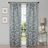 Two light gray curtains with a leaf pattern hang in front of a window. They are accompanied by framed floral artwork on the wall and a lamp on a small decorative table.