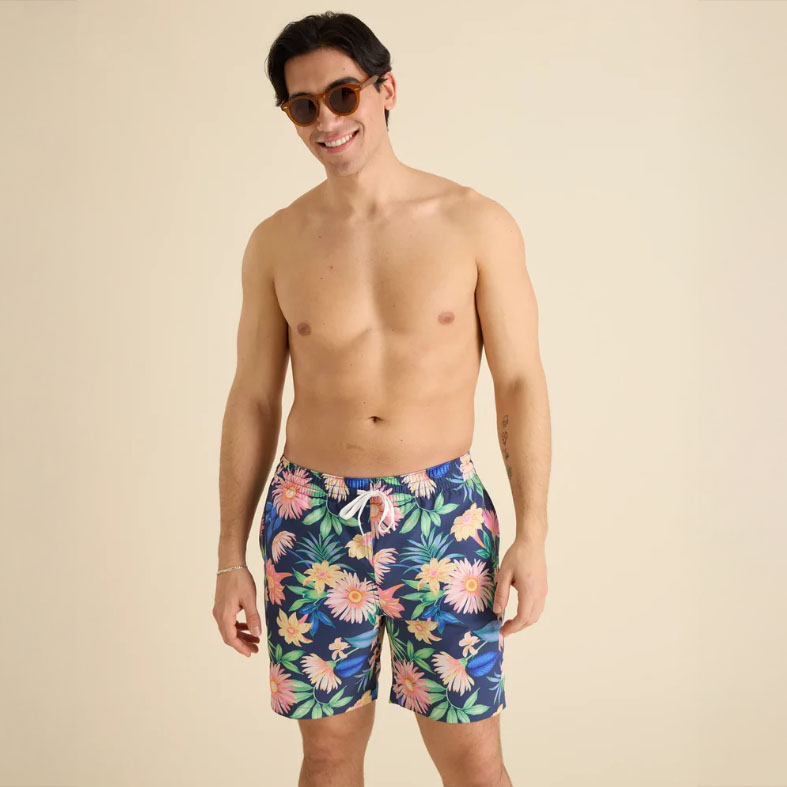 A man is wearing floral-patterned swim shorts with a navy background and elastic waistband, paired with round sunglasses.