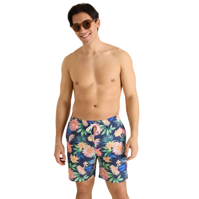 A man is wearing floral-patterned swim shorts with a navy background and elastic waistband, paired with round sunglasses.