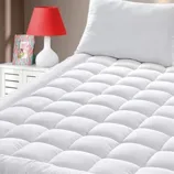 A plush, quilted mattress topper on a bed with a white pillow, accompanied by a red lamp and a book on a nightstand.