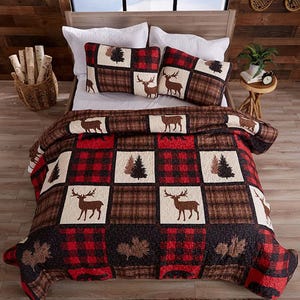 Quilt set featuring a checkered pattern with deer and tree designs in red, brown, and white, shown on a bed with matching pillows and a rustic decor setting.