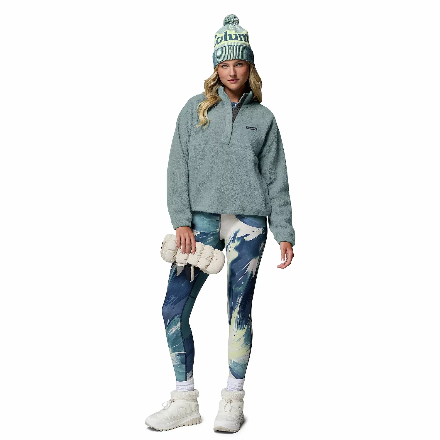 A person is wearing a light blue fleece pullover, abstract-pattern leggings, white boots, and holding a cream-colored puffer bag. They have a beanie with the word \“Columbia\“ and a pom-pom on top.