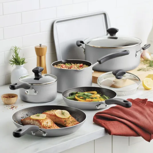 A set of nonstick cookware, including two frying pans, two pots with lids, and a large pot with a lid, all in a light gray color. The set is displayed with cooked salmon, vegetables, and quinoa, along with kitchen accessories.