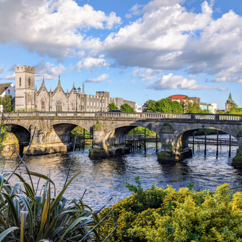 Stone arch bridge over a river with a church and buildings in the background, surrounded by greenery and yellow flowers under a partly cloudy sky.