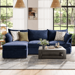 Blue sectional sofa with mixed-pattern pillows, gray coffee table with decor, large windows, hanging lamps, abstract wall art, and patterned rug on wooden floor.