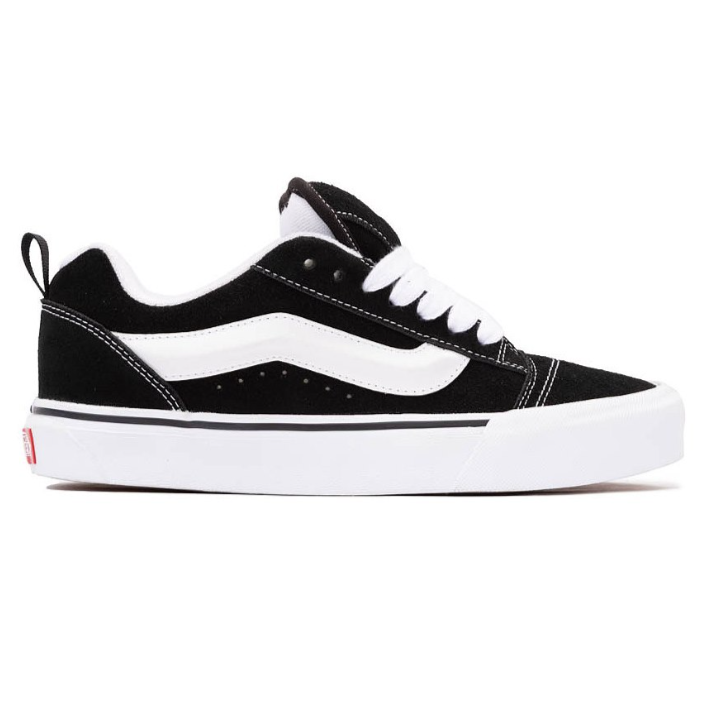 A black and white low-top sneaker with a suede upper, featuring a white side stripe, white laces, and a white sole with a small red detail on the heel.