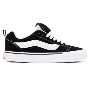 A black and white low-top sneaker with a suede upper, featuring a white side stripe, white laces, and a white sole with a small red detail on the heel.