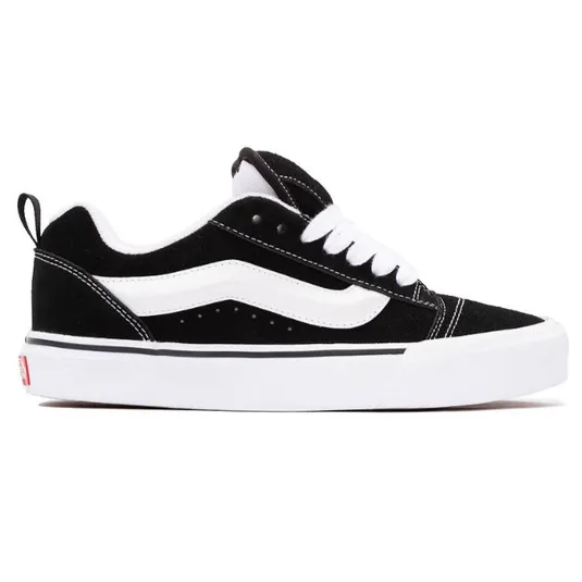 A black and white low-top sneaker with a suede upper, featuring a white side stripe, white laces, and a white sole with a small red detail on the heel.