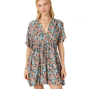 A woman wears a floral-patterned dress with short, loose sleeves and a V-neckline, featuring shades of pink, blue, and green. She also has a necklace and a bracelet.