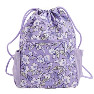A purple Vera Bradley Drawstring Backpack features a floral pattern with white and blue flowers, a front zipper pocket, side pockets, and matching purple drawstrings.