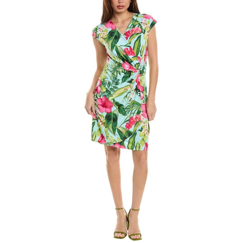 A woman is wearing a knee-length, short-sleeved dress with a vibrant tropical floral pattern in green, pink, and white, paired with green strappy sandals.