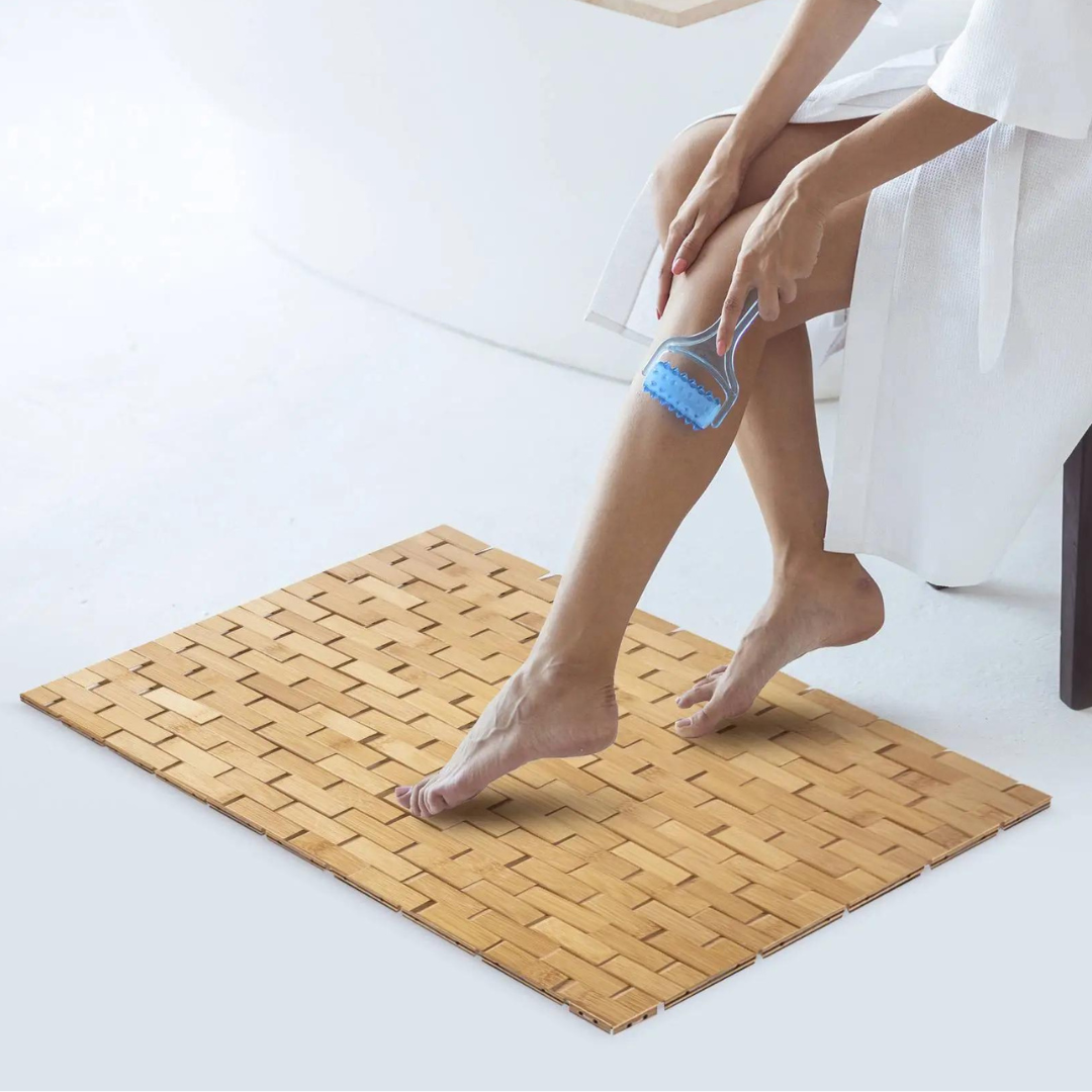 A person uses a blue loofah on their leg while standing on a woven bamboo mat.