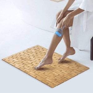 A person uses a blue loofah on their leg while standing on a woven bamboo mat.