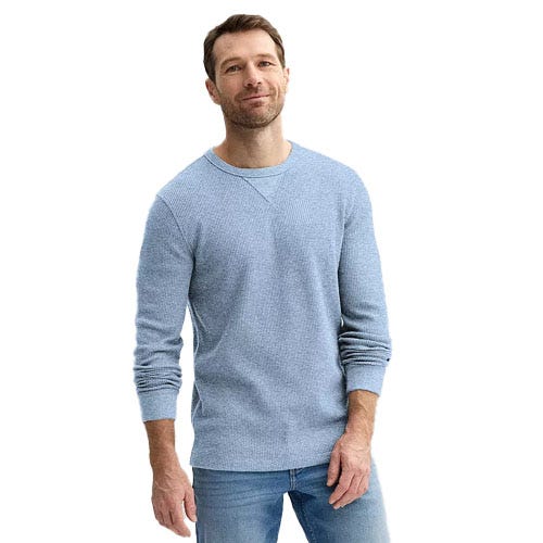 A man is wearing a light blue long-sleeve sweater and light blue jeans, standing with a slight smile.