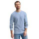 A man is wearing a light blue long-sleeve sweater and light blue jeans, standing with a slight smile.