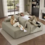 Two people relax on a large, ribbed beige sectional sofa in a living room featuring a fireplace, bookshelf, and large window with curtains.