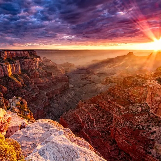A breathtaking view of the Grand Canyon during sunset, showcasing the vivid colors and rugged terrain under a dramatic sky with sun rays streaming through the clouds.