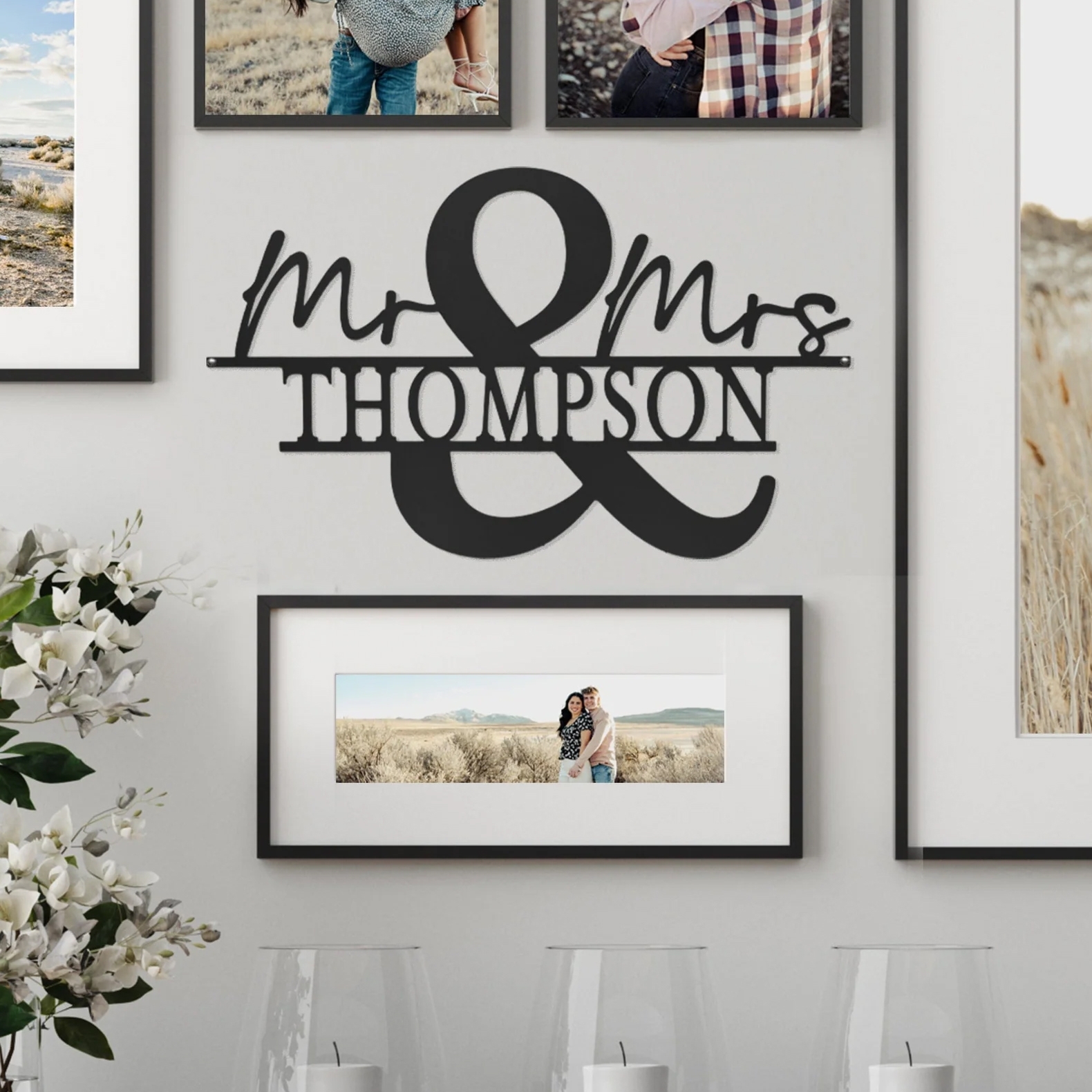 Wall art displays the text \“Mr & Mrs Thompson\“ in black, surrounded by framed photographs. White flowers and candles are positioned below the frames.