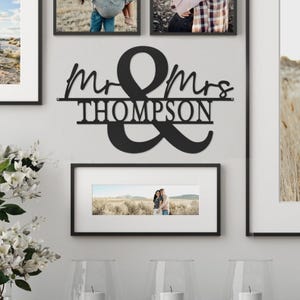 Wall art displays the text \“Mr & Mrs Thompson\“ in black, surrounded by framed photographs. White flowers and candles are positioned below the frames.