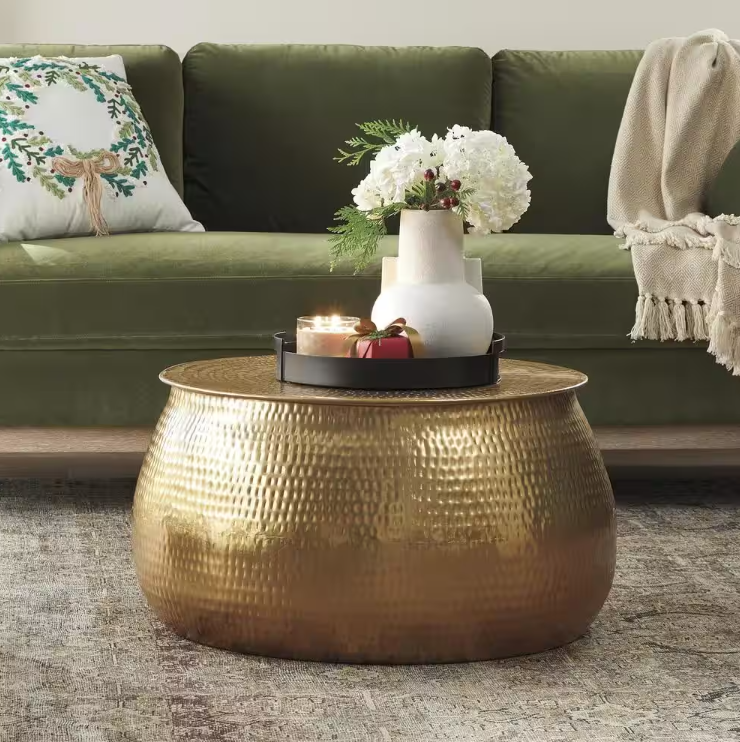 A hammered gold coffee table is topped with a black tray holding a lit candle and a white vase with white flowers. The setting includes a green sofa with a festive pillow and a beige throw blanket.