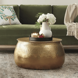 A hammered gold coffee table is topped with a black tray holding a lit candle and a white vase with white flowers. The setting includes a green sofa with a festive pillow and a beige throw blanket.