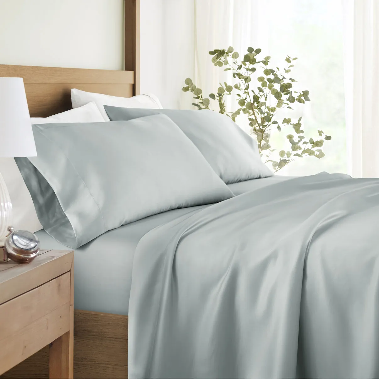 A neatly made bed featuring light gray silk sheets and pillowcases, set against a wooden headboard with a plant and a bedside table with a lamp and a clock.