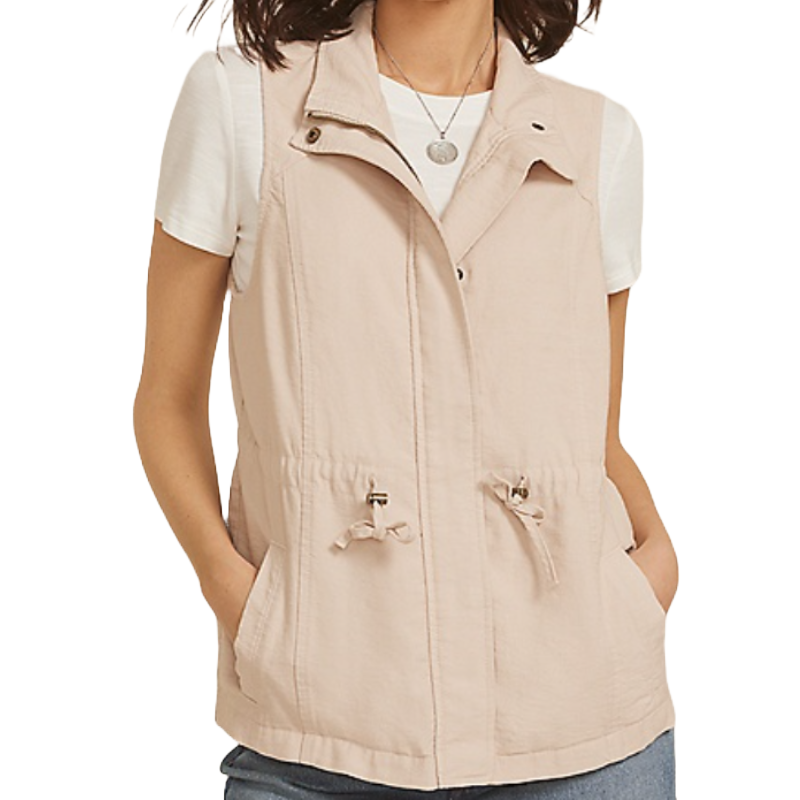 A woman is wearing a sleeveless utility vest in a light beige color, paired with a white t-shirt and blue jeans. The vest features a stand-up collar, zip closure, and waist drawstrings.