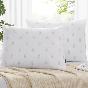 Two white pillows with a subtle anchor and \“N\“ pattern are placed on a bed with a textured beige blanket and an open book nearby.