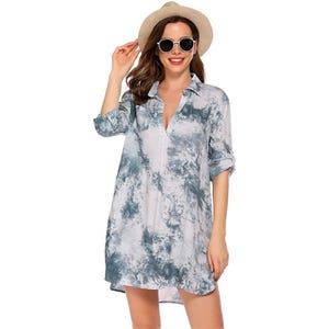 A woman is wearing a tie-dye swim cover-up dress with rolled-up sleeves, featuring a V-neck and a loose, flowy fit. She accessorizes with a straw hat and round sunglasses.