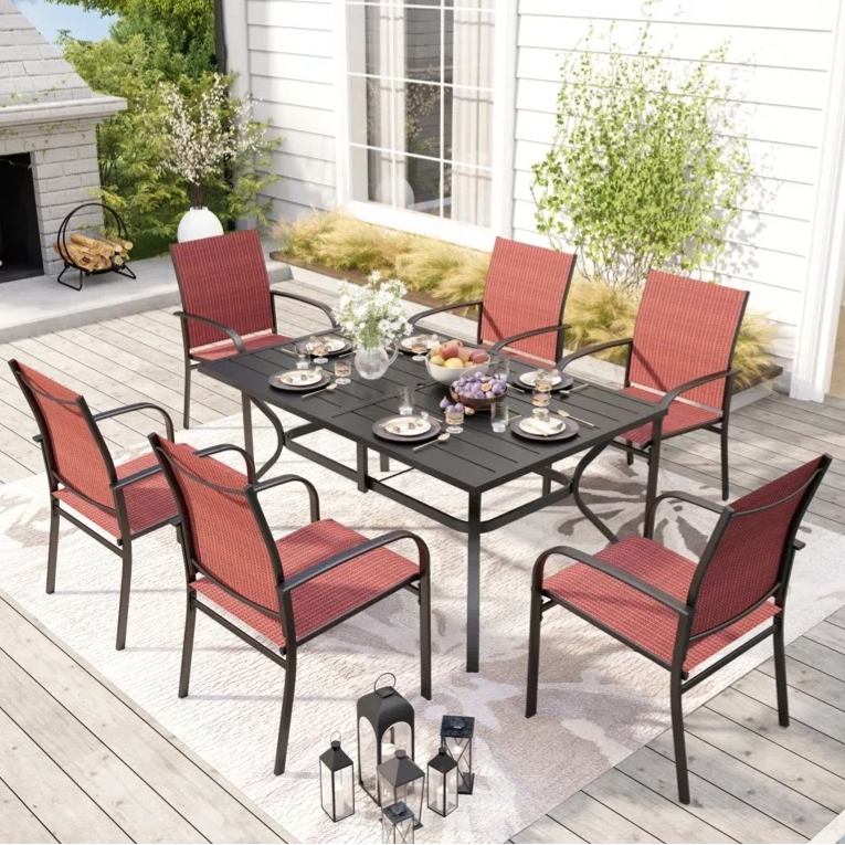 A patio dining set featuring a black table and six red cushioned chairs is arranged on a patterned outdoor rug, accented by black lanterns and surrounded by greenery and a fireplace.