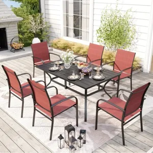 A patio dining set featuring a black table and six red cushioned chairs is arranged on a patterned outdoor rug, accented by black lanterns and surrounded by greenery and a fireplace.