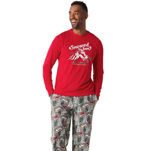 The North Pole Men's 2pc Matching PJ Set features a red long-sleeve top with \“Snowed Inn\“ and skiing graphic, paired with gray pants adorned with a festive tree pattern.