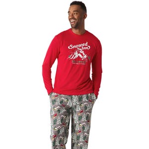 The North Pole Men's 2pc Matching PJ Set features a red long-sleeve top with \“Snowed Inn\“ and skiing graphic, paired with gray pants adorned with a festive tree pattern.