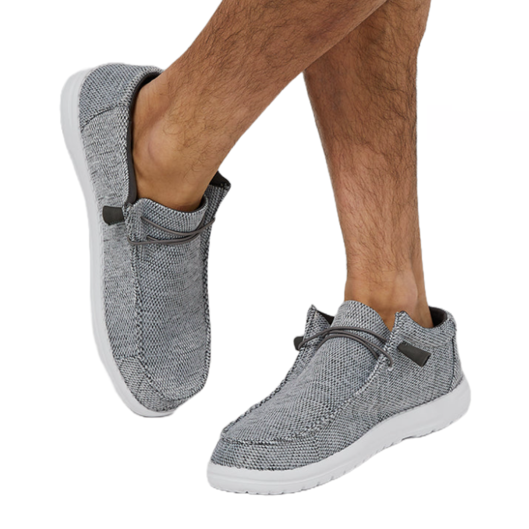 Casual slip-on shoes with a grey textured fabric, white soles, and decorative laces, worn by a person with bare legs.