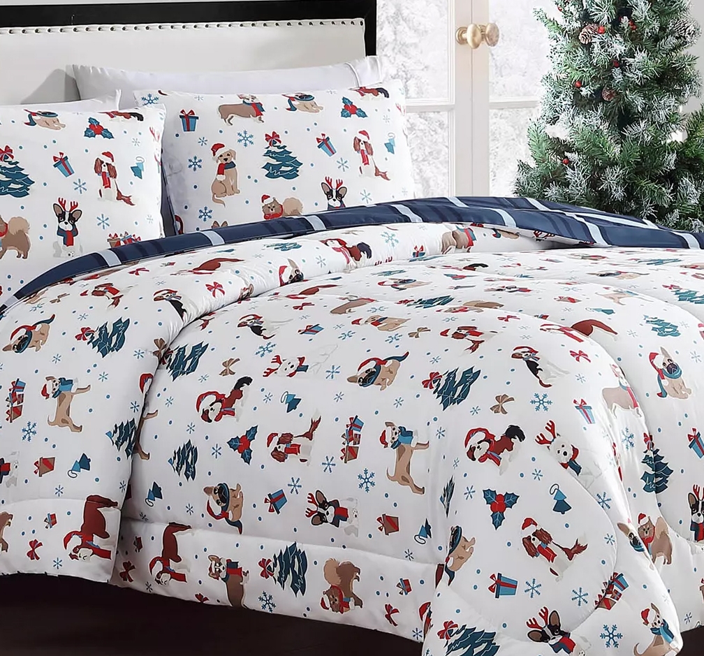 Festive bedding set with a holiday theme, featuring illustrations of dogs, Christmas trees, gifts, and snowflakes in red, blue, and brown on white fabric, complemented by navy striped sheets.