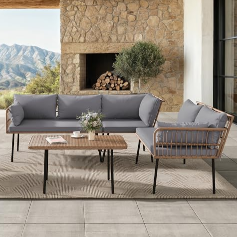 Outdoor patio set with a stone backdrop, including a grey cushioned sofa, two chairs, and a rectangular wooden coffee table with metal legs, placed on tiled flooring. A small plant and fireplace with stacked wood complete the scene.