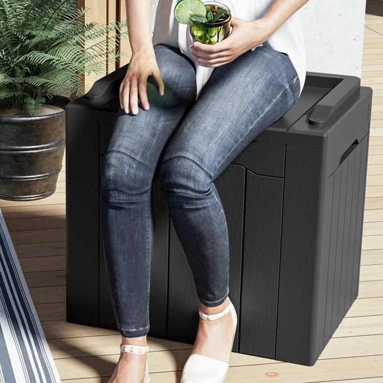 Person sitting on a sturdy outdoor storage box that doubles as a seat, with a maximum capacity of 220 lbs. The setting includes a rug, a couch with a green pillow, and a potted plant.