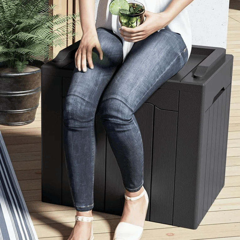 Person sitting on a sturdy outdoor storage box that doubles as a seat, with a maximum capacity of 220 lbs. The setting includes a rug, a couch with a green pillow, and a potted plant.