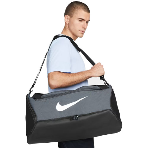 A person is carrying a large Nike gym duffel bag featuring a prominent white swoosh logo, adjustable shoulder strap, and a combination of gray and black fabric.