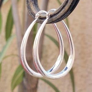 A pair of silver teardrop-shaped hoop earrings is hanging on a metal rod, set against a blurred outdoor background.