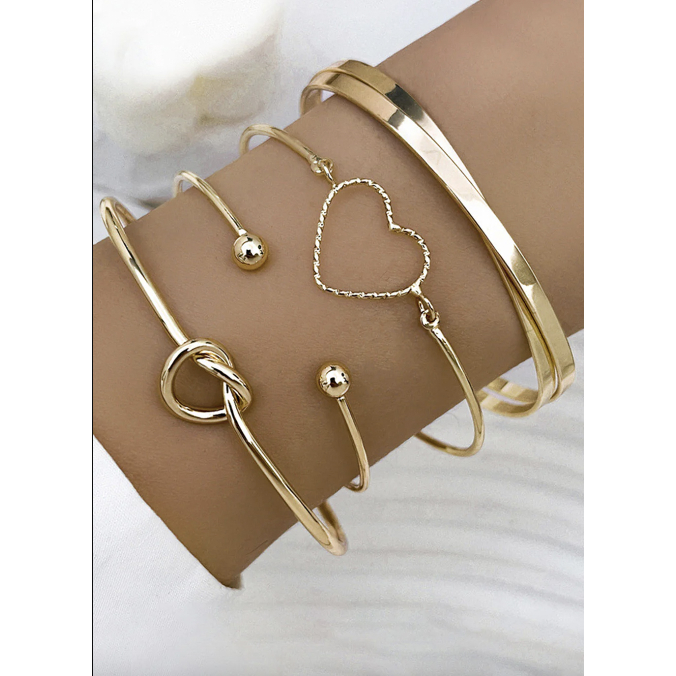 A 4-piece bracelet set featuring gold-tone designs: a solid polished bangle, a heart-shaped open wire bracelet, a twisted knot bracelet, and an open bracelet with round ends.