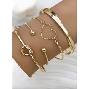 A 4-piece bracelet set featuring gold-tone designs: a solid polished bangle, a heart-shaped open wire bracelet, a twisted knot bracelet, and an open bracelet with round ends.