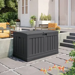 A black outdoor storage deck box is placed on a tiled patio with surrounding outdoor furniture and plants.