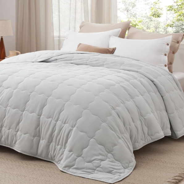 A light gray, quilted down-alternative comforter is displayed on a bed with white and brown pillows, against a backdrop of sheer curtains. The comforter features a soft texture and a subtle geometric pattern.