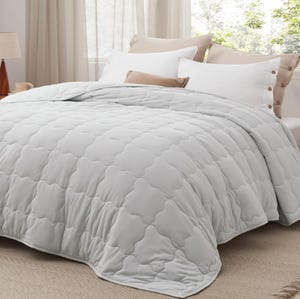 A light gray, quilted down-alternative comforter is displayed on a bed with white and brown pillows, against a backdrop of sheer curtains. The comforter features a soft texture and a subtle geometric pattern.