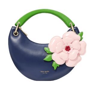 A Kate Spade New York handbag featuring a blue crescent shape with a green handle and adorned with a large pink flower with green leaves on the front.